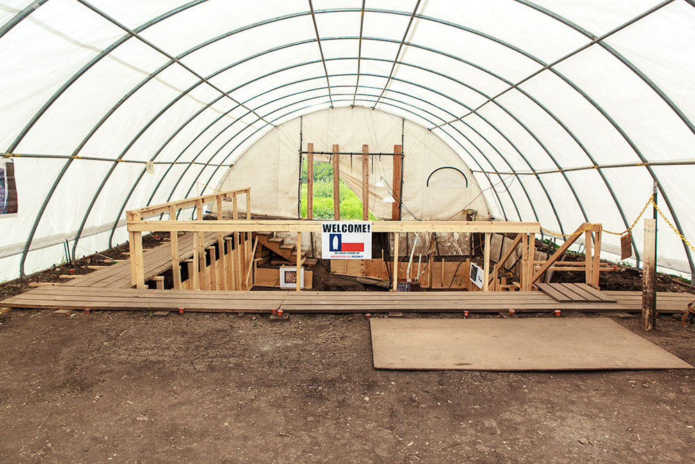 inside of white tent with welcome sign attached to wooden platforms leading down into the Gault dig site.