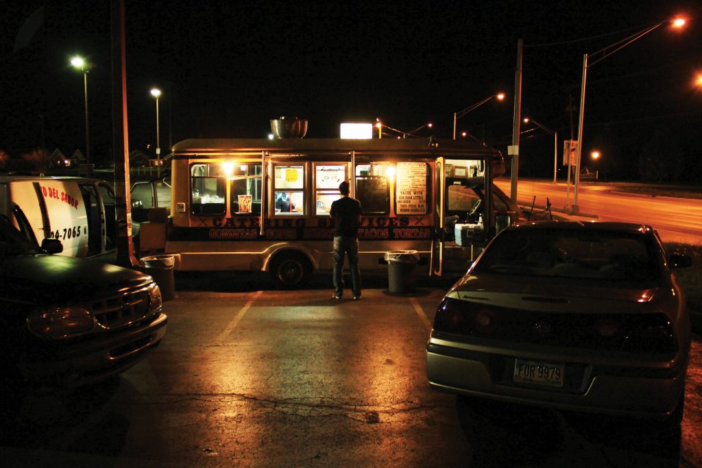 A man standing outside a taco truck at night.