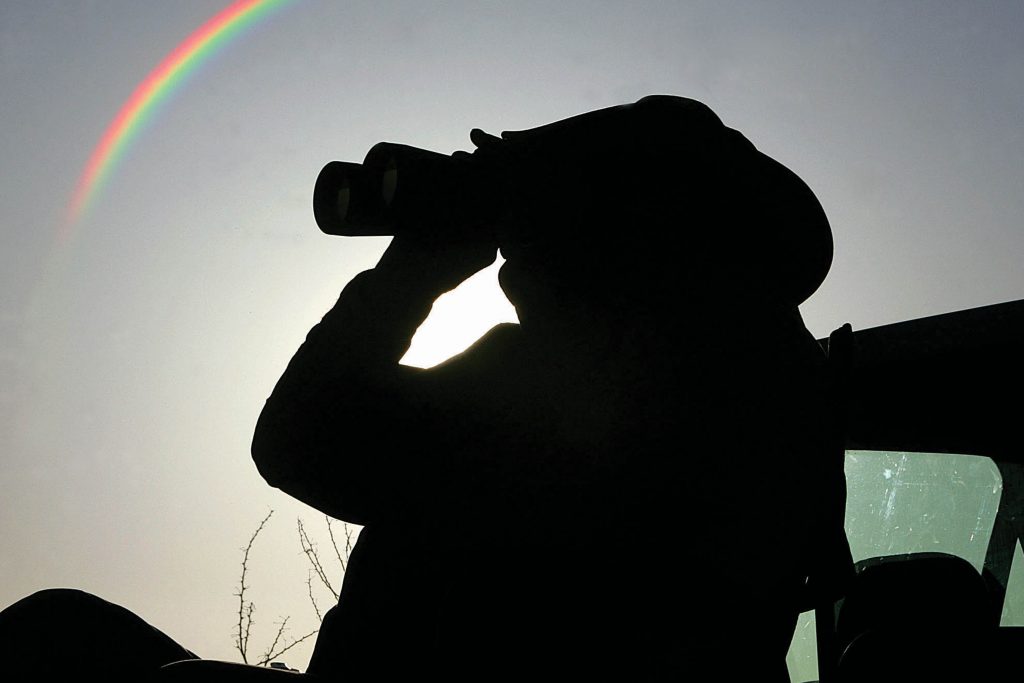 A silhouette of a Minuteman wearing a cowboy hat looking through binoculars. A rainbow is in the clear sky behind him.