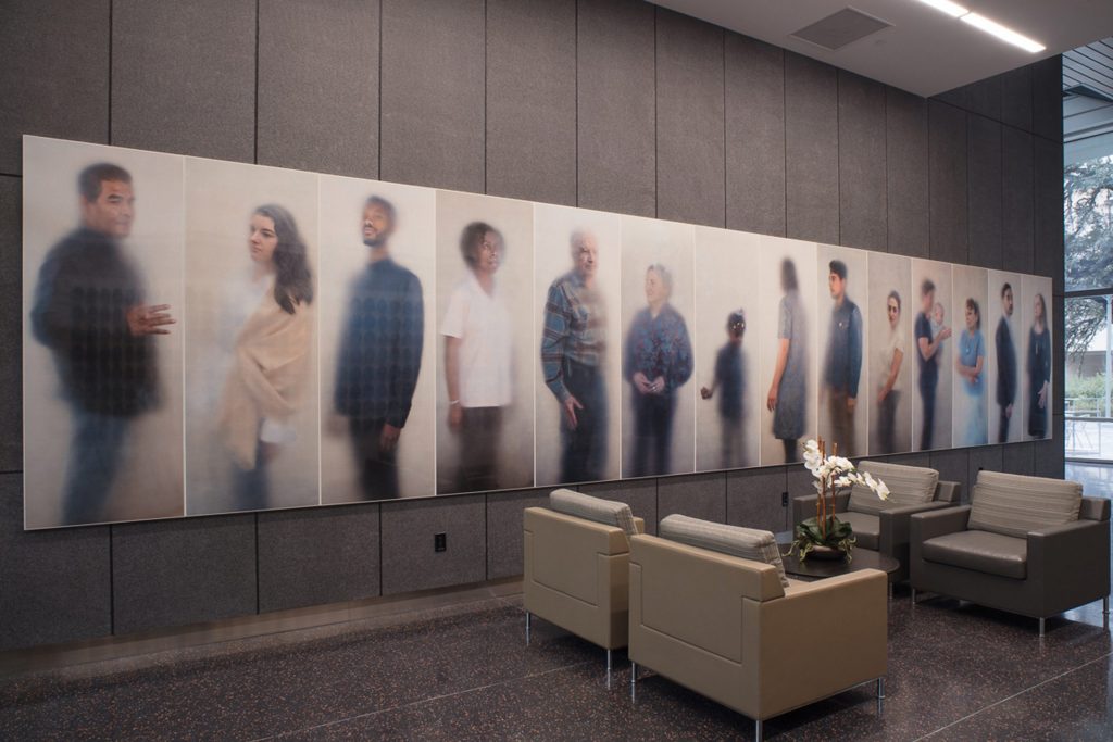 Photo of the lobby of the Dell Medical School. A series of 14 portraits from Ann Hamilton's O N E E V E R Y O N E series hang on the main wall to the entrance.