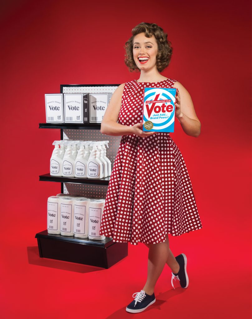 A tongue-in-cheek photo of a smiling woman holding a brightly branded "Vote" box against a red background. The box reads, "The Campaign for Your Vote: Just Add Brand Power!" (America's Choice). She is steping towards the camera and behind her is a supermarket shelf of "Generic Branded" black and white boxes.