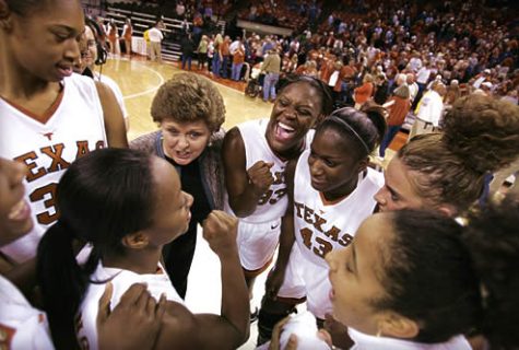 A group of women college basketball players and their coach in a huddle with the court and fans in the background