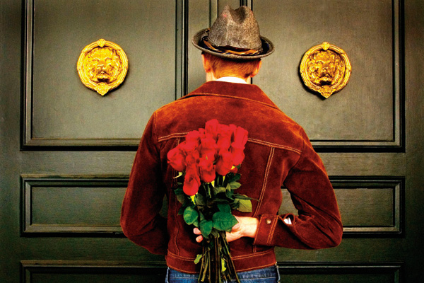 man in hat and red jacket waiting at door with roses behind his back