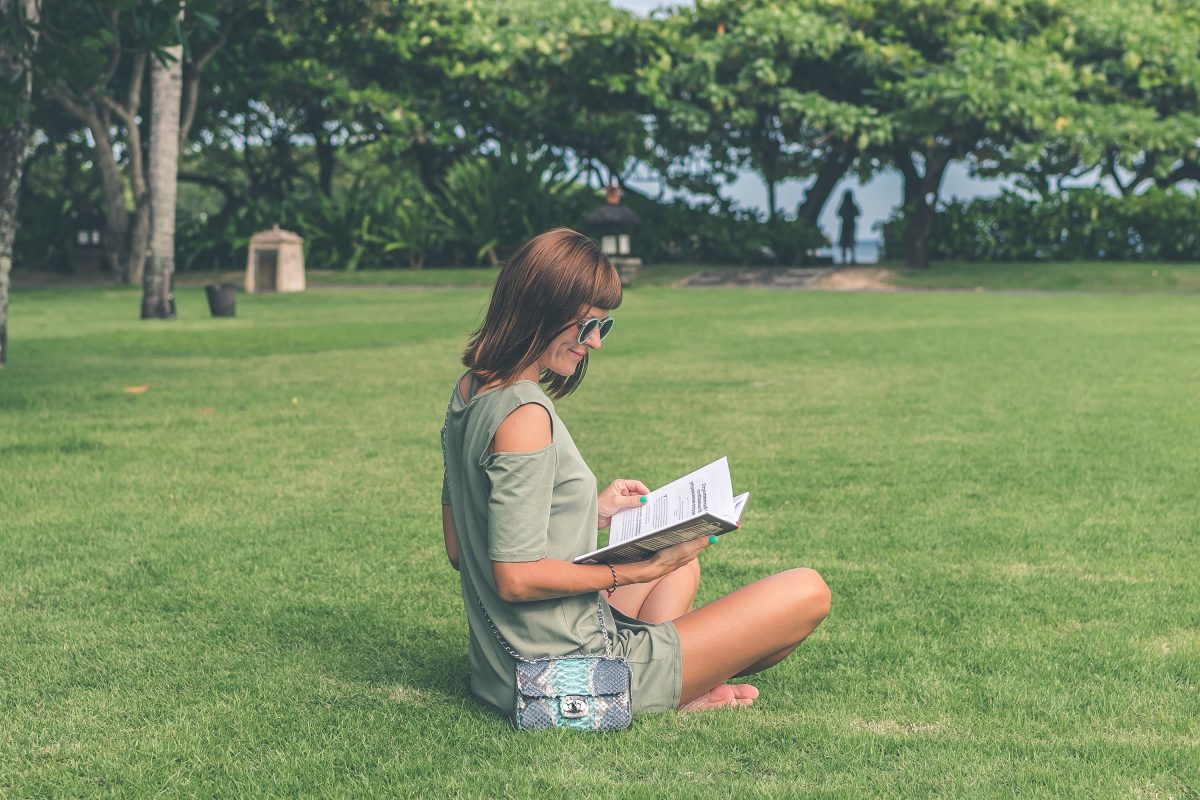 Woman with sunglasses sitting on grass reading book.
