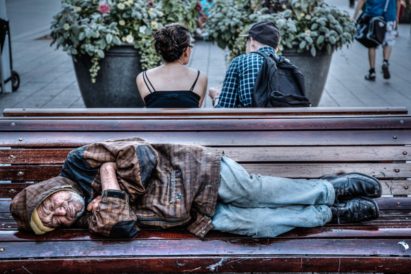 homeless man sleeping on bench opposite a couple