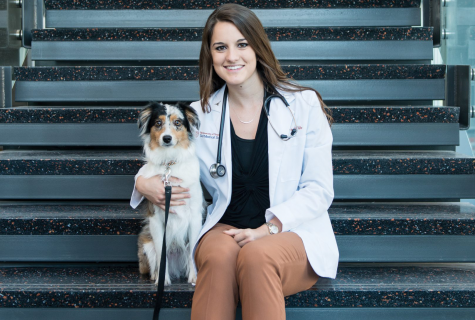 Cierra Grubbs sits on the stairs of Dell Medical School with her arm around her dog. She's wearing a lab coat and stethoscope.