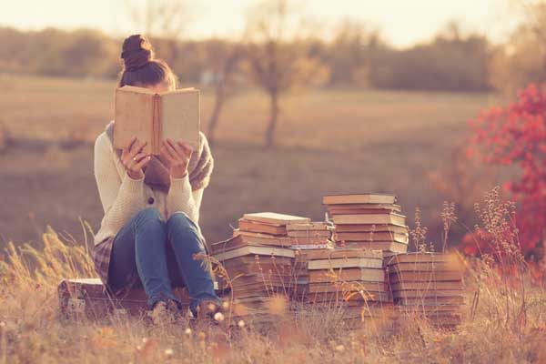 Woman reading a book while sitting on suitcase next to stack of books outdoors with fall colors.
