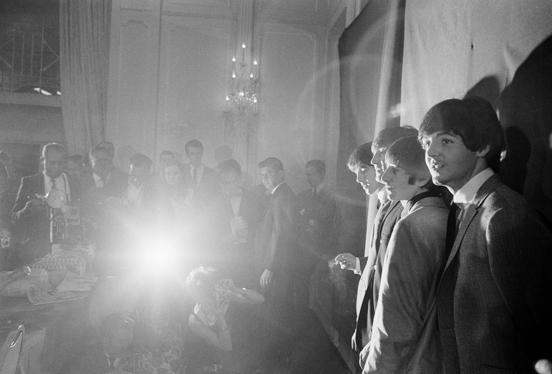 Black and white photograph of The Beatles standing against a white backdrop as a crowd looks on.