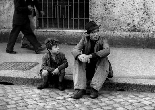Black and white photo of a man and young boy sitting on a sidewalk.