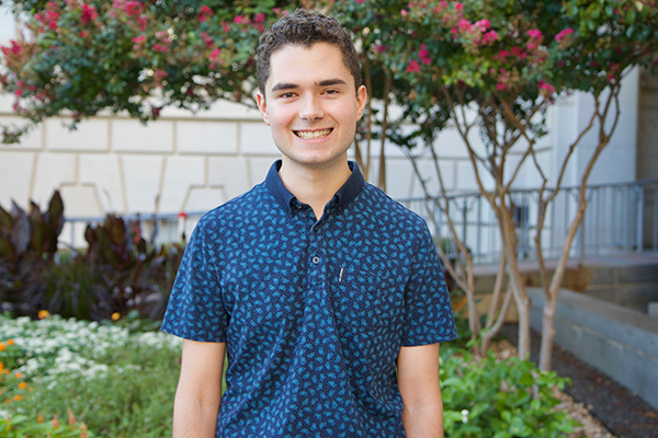 Isaac James smiles happily in front of a flower bed with a variety of plants, wearing a patterned blue shirt.