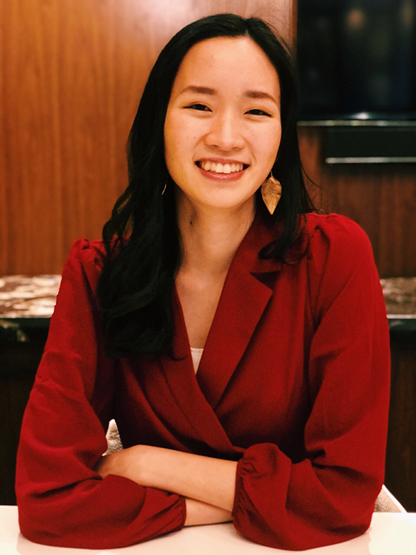 Margaret Siu smiles at the camera. She is wearing a flowy red top and metal earrings carved in the shape of a leaf. Her arms are crossed on the table below her.