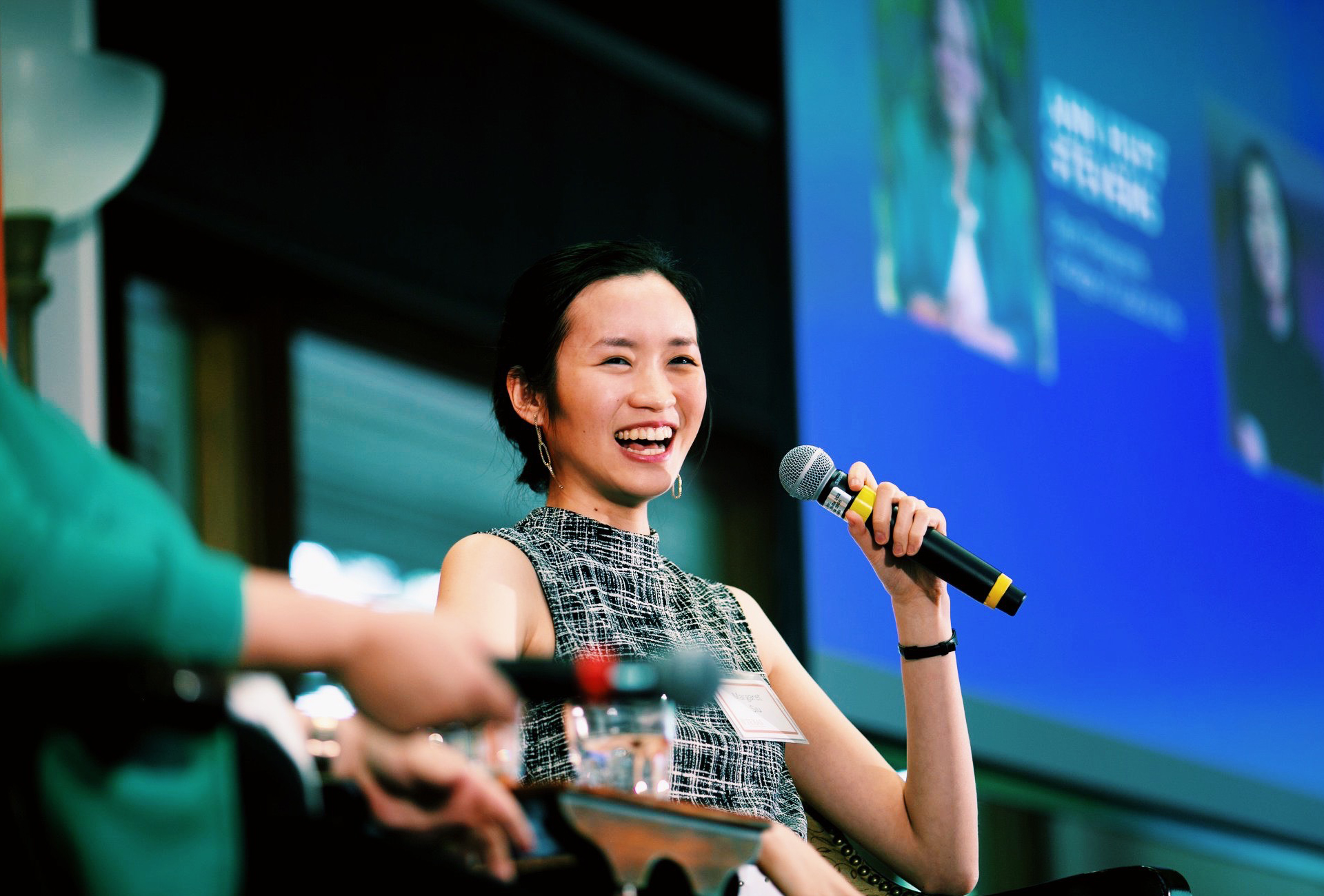 Margaret Siu sits in a chair while holding a microphone up to her mouth. She is laughing happily at what is clearly a public speaking event.