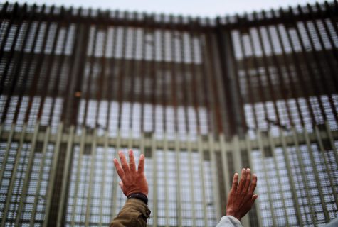 Photo of two outstretched hands in front of a large, imposing border fence.