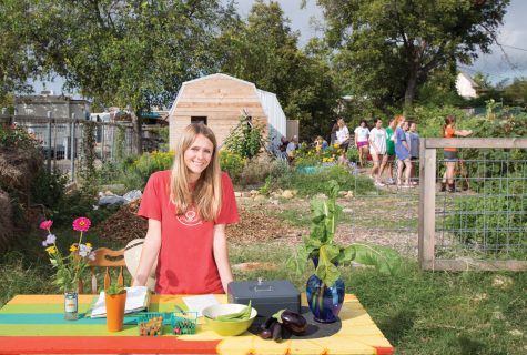 Photo of community garden with a smiling woman at the vegetable stand. A large shed is in the background along with a group of students being shown how to harvest vegetables.