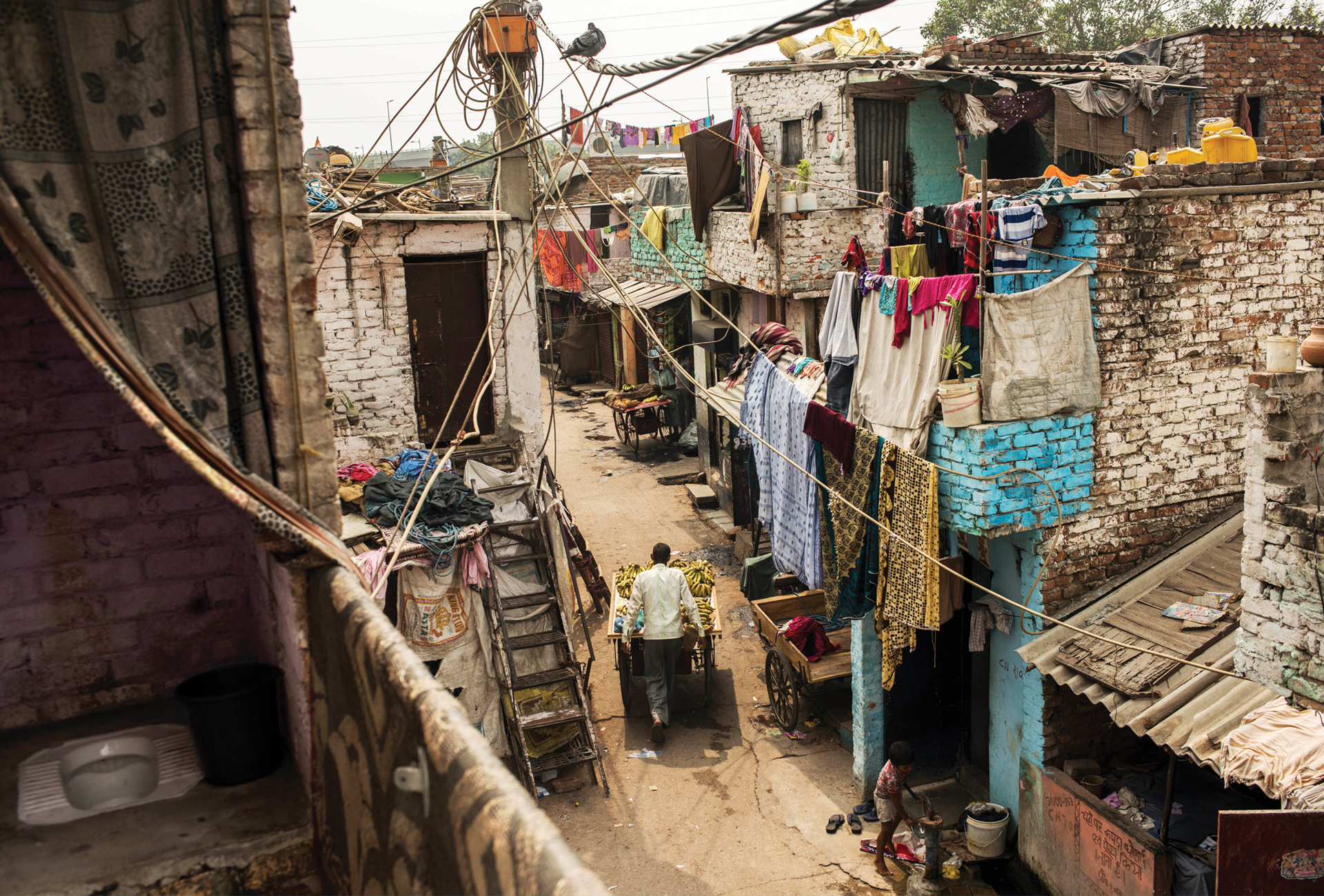 Photo from a balcony in Safeda Basti, India. The street scene below appears dirty and is crowded with laundry hanging from clotheslines while a lone man pushes a banana cart. To the immediate left, we see an open-air squat toilet overlooking a balcony.