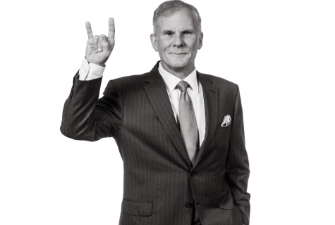 Stuart Stedman stands with one hand in his pocket and the other raised in a Hook Em sign. He is wearing a suit and tie.