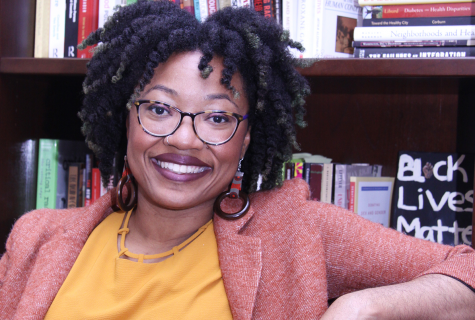 Ashanté Reese sits in front of a full book shelf. She is smiling widely and wearing a red blazer and light orange blouse.