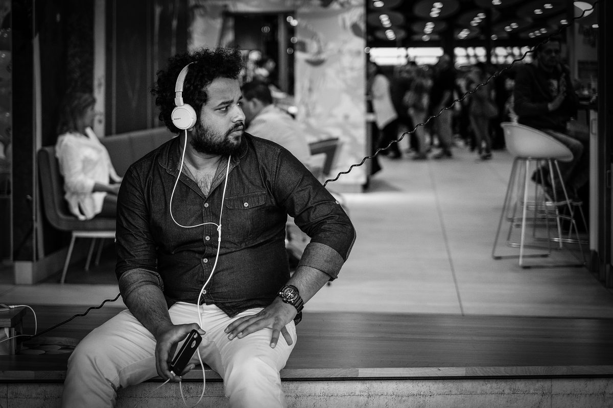 A man sits on a bench outside, listening to earbuds plugged into his phone and looking off to his left.