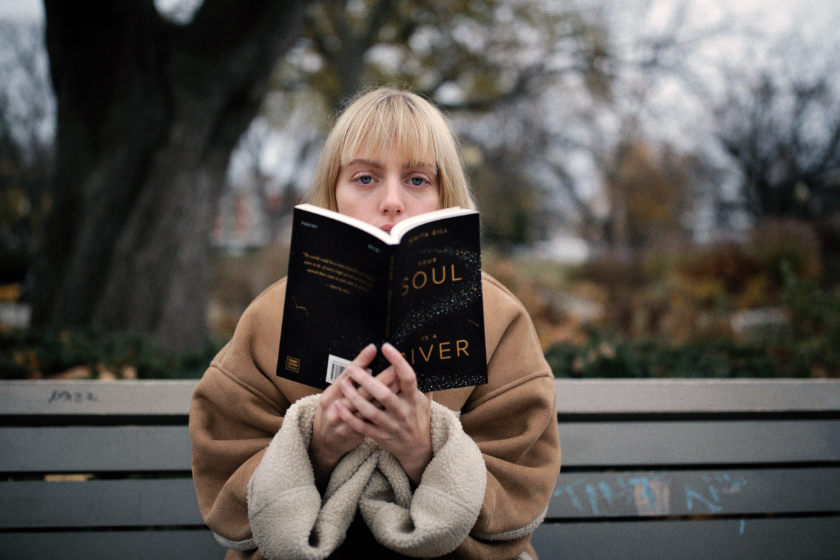 A woman with a blonde bob sits on a bench wearing a warm coat and reading a book she has held out in front of her, hiding the bottom half of her face.