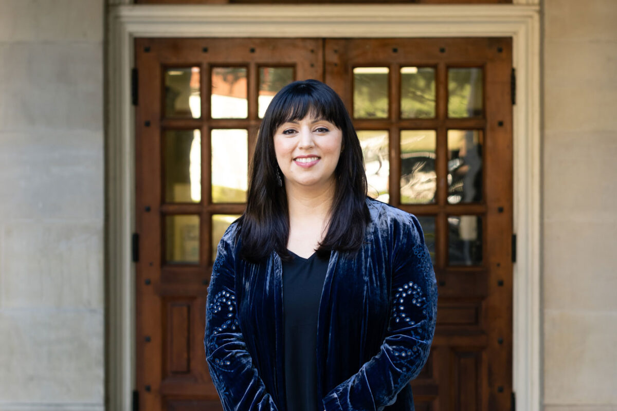 Monica Muñoz Martinez stands on campus in front of two wooden doors. She is smiling widely and wearing a blue velvet blazer.