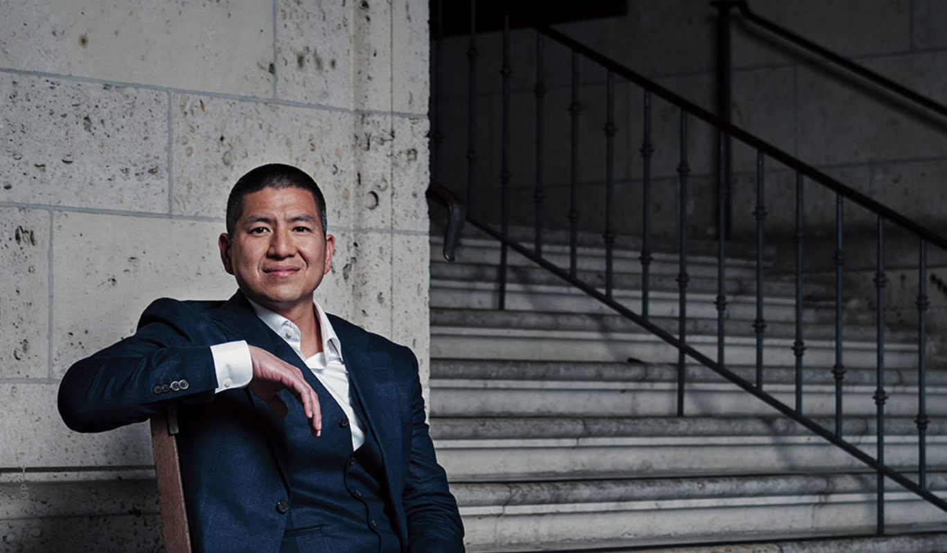 Eric Tang is sitting in a chair and dressed in a blue suit with an open collar. Behind him is a stone staircase on the UT campus.