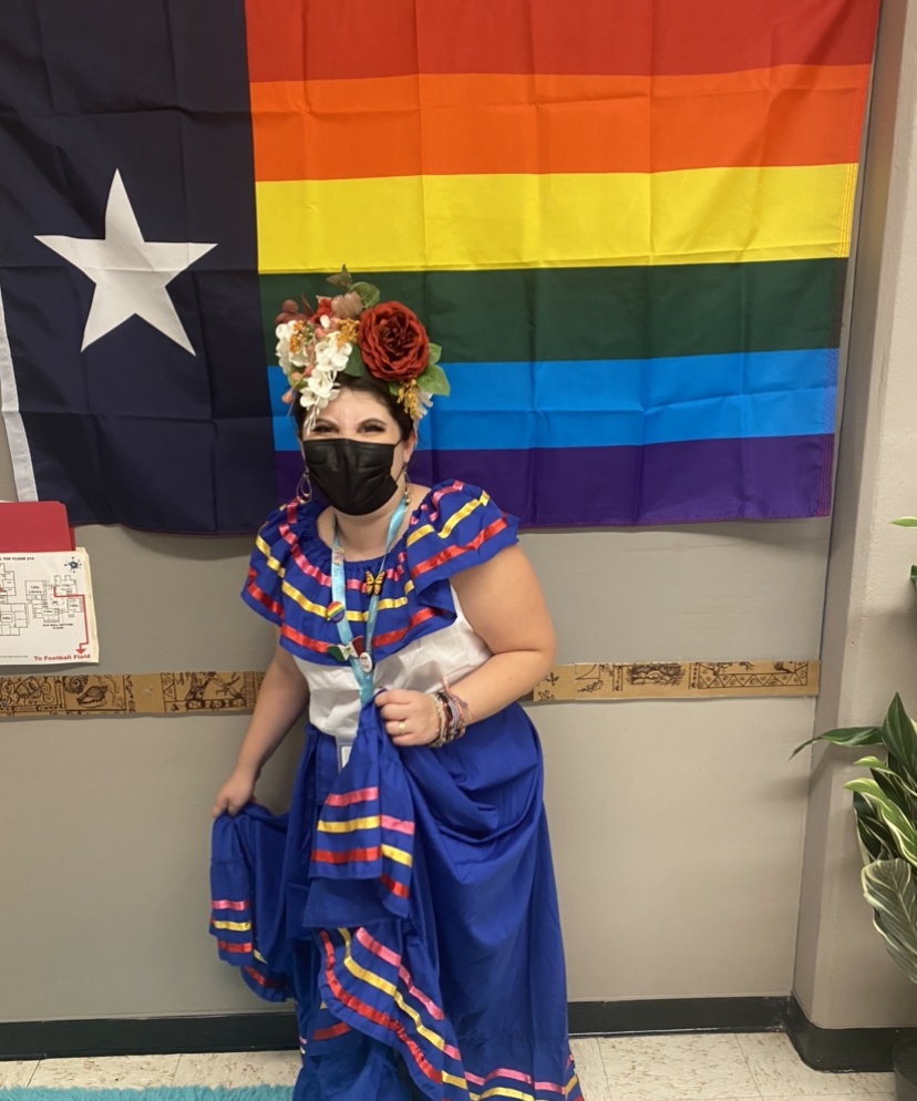 Yulissa wearing traditional Mexican clothing with an elaborate flower crown on her head. She's wearing a black mask and standing in front of an LGBT Texas flag.