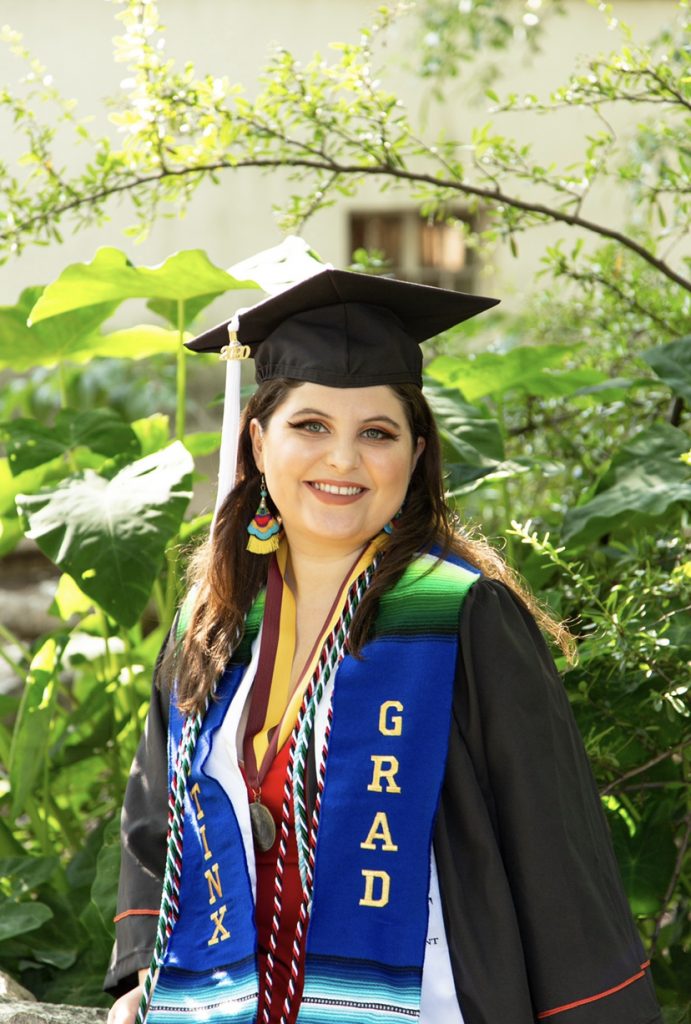 Yulissa on campus in front of greenery wearing her graduation cap, robers and a Latinx Grad stole.