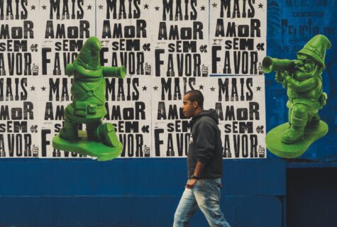 Brazilian man in hoodie walks to the left in front of wall plastered with posters reading Mais Amor Sem Favor with superimposed green figures